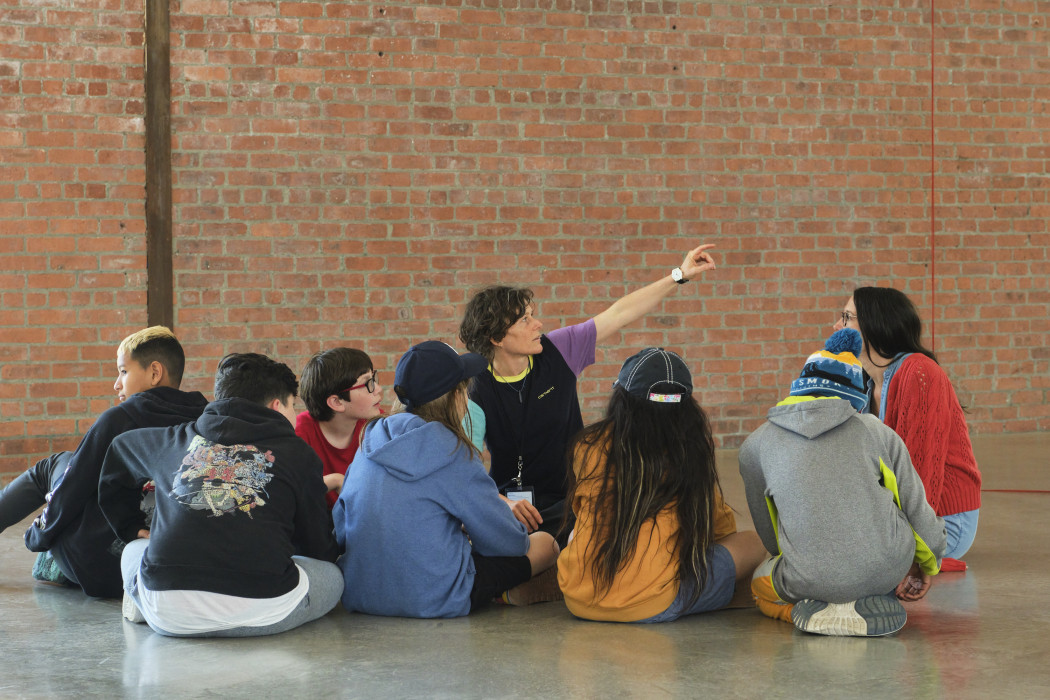 A group of young kids and teens sit on the concrete floor around an adult teacher who is pointing up to the right. Behind them is a brick wall.